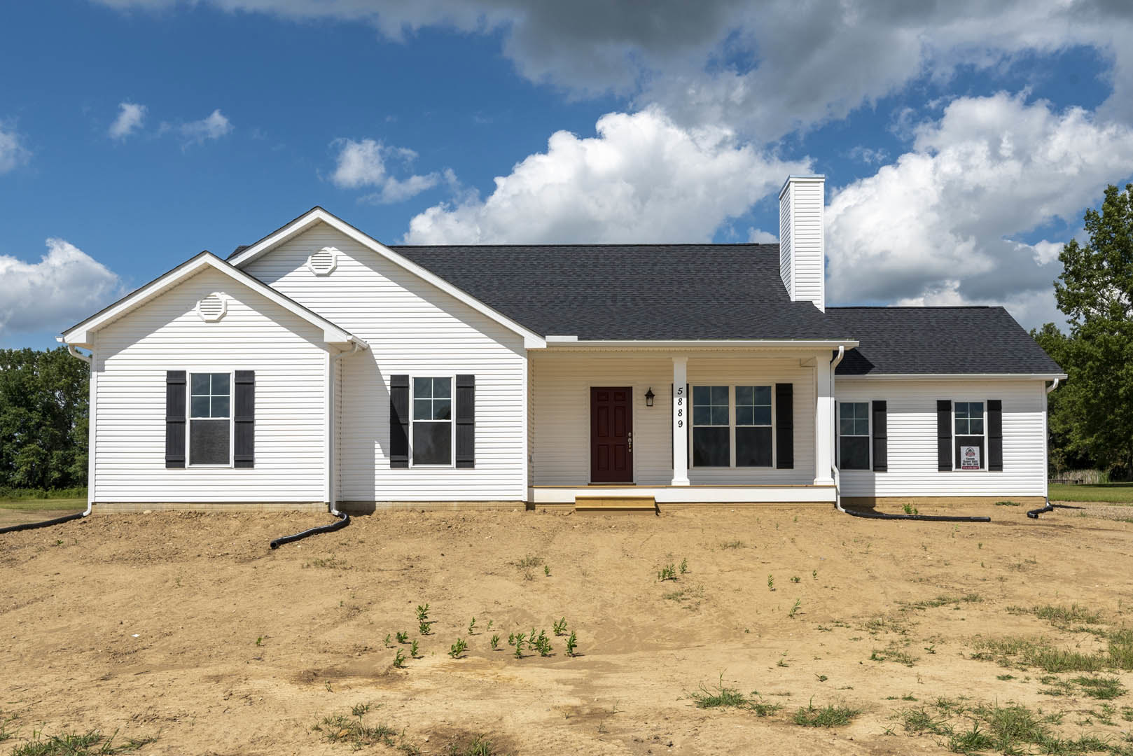White house with black shutters, multiple windows, and a dirt yard with sparse plants on a sloped hill