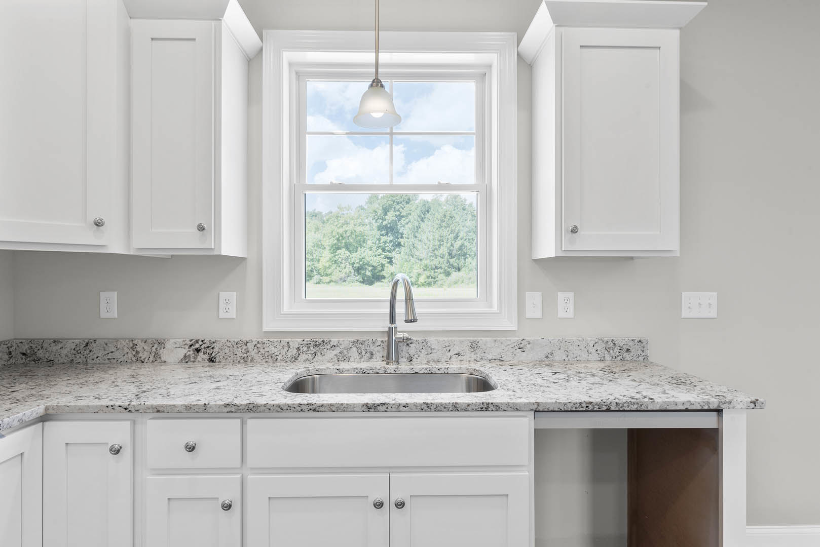 White shaker cabinets and drawers, stainless steel sink with chrome faucet, quartz countertop, rectangular electrical outlet, pendant light fixture, window with natural light.