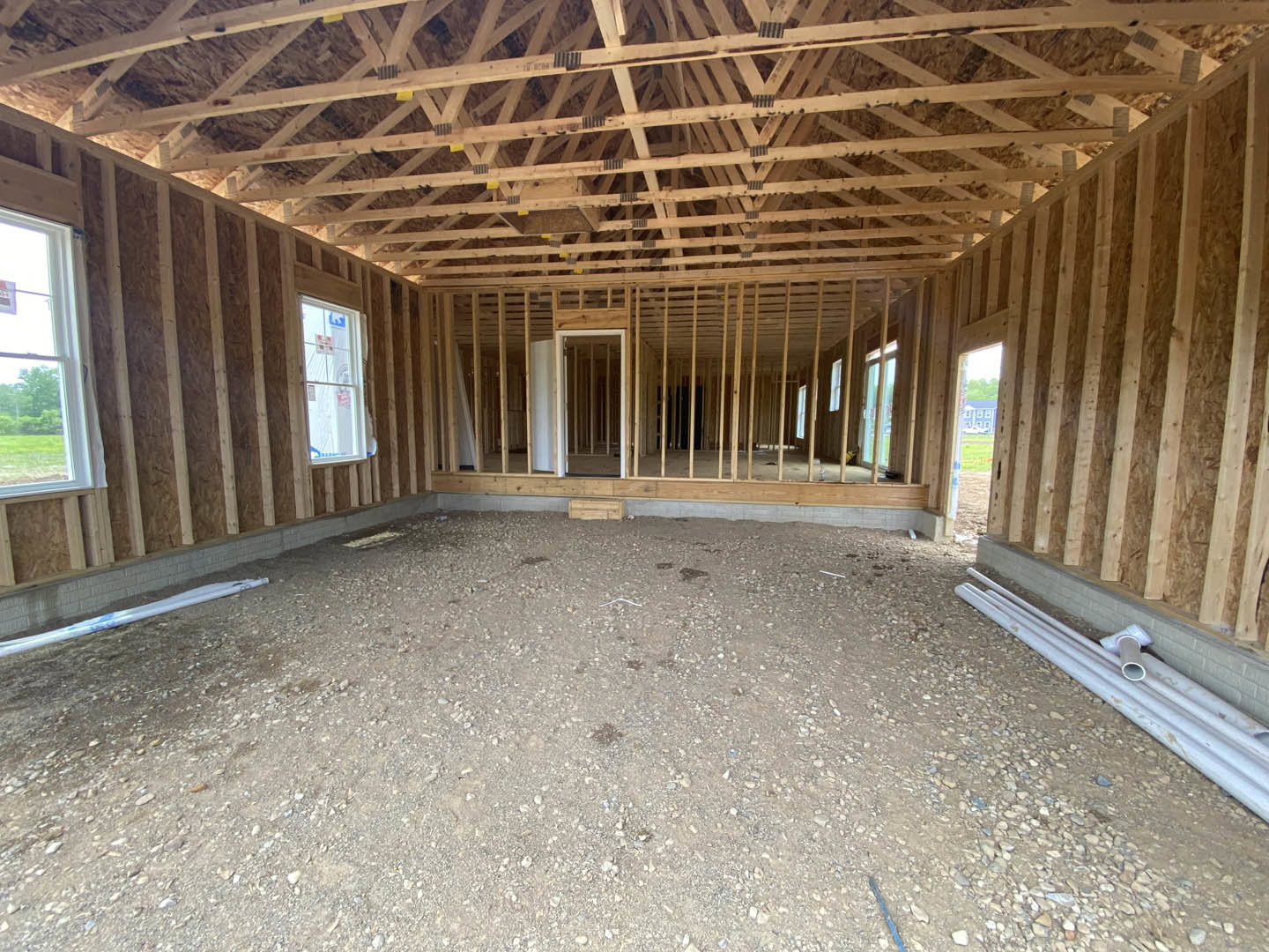 Unfinished residential interior with exposed wooden ceiling beams, gravel and rocks covering the dirt floor, white-framed window, close-up of a door, and white plumbing pipes