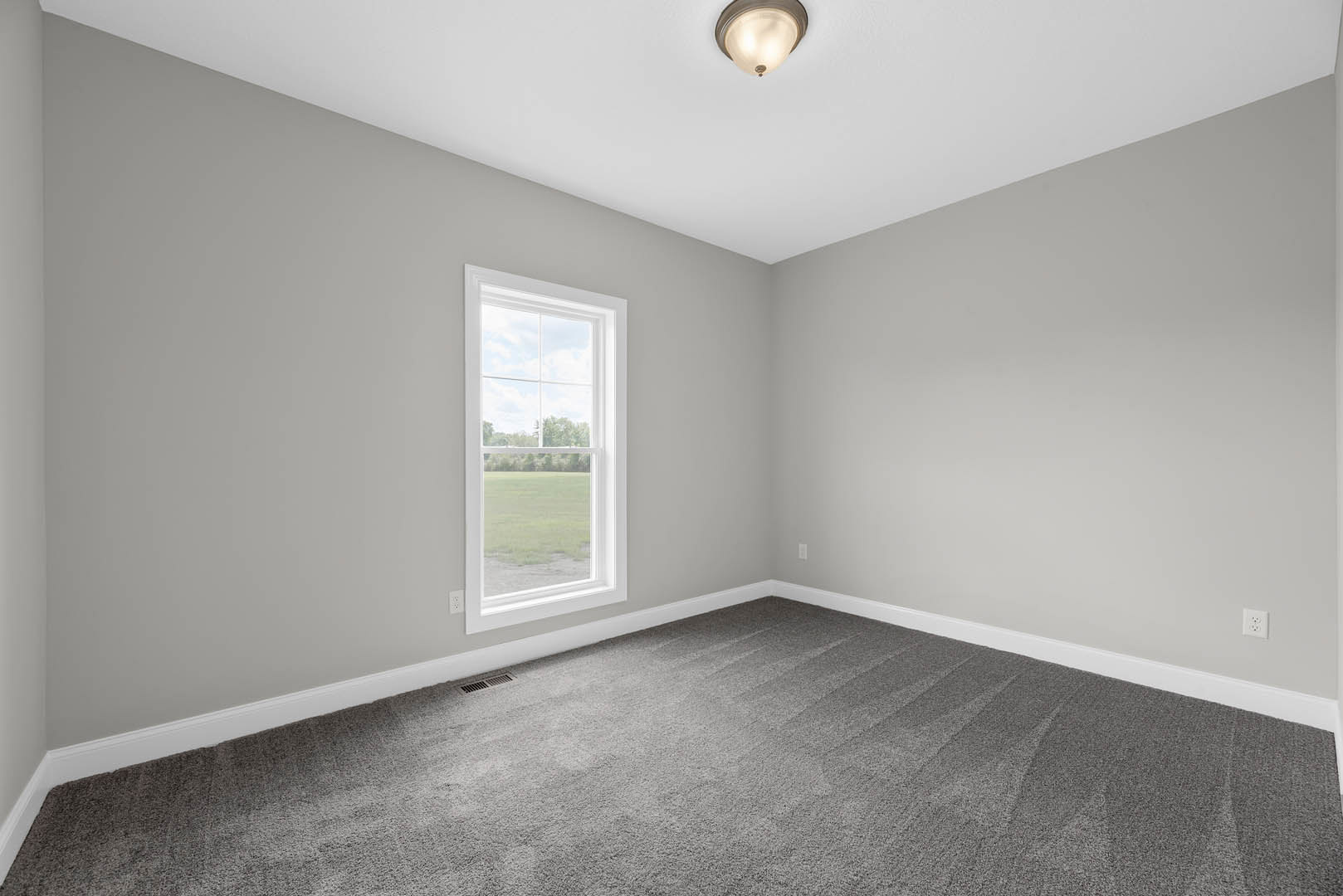 Bright room with white walls, grey carpet flooring, large window overlooking a green field, and modern ceiling light fixture