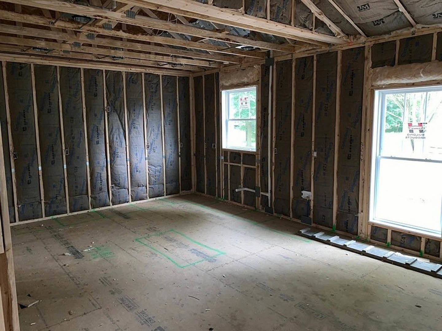 Open room featuring exposed wood beams, large windows, unfinished wall with black paper, green line marking on floor, and wooden ceiling intersected by a metal beam.