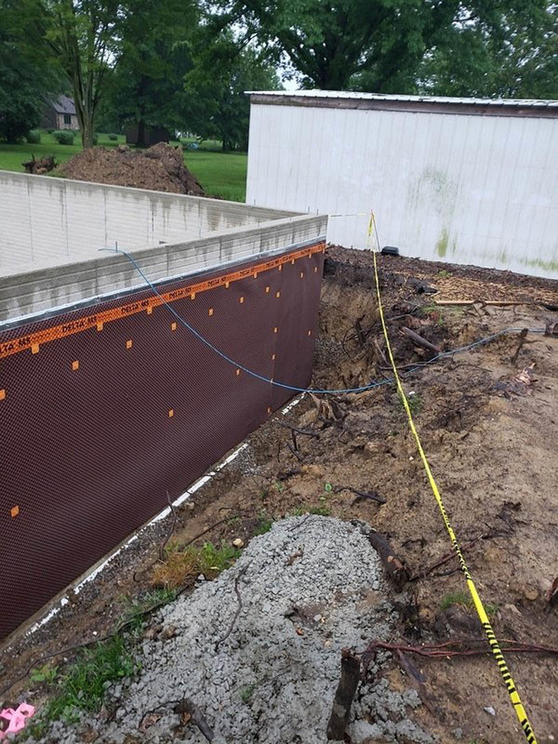 White concrete wall with yellow caution tape, pile of gravel and grass, blue rope, exposed dirt and hole in ground, blurred pink object in foreground