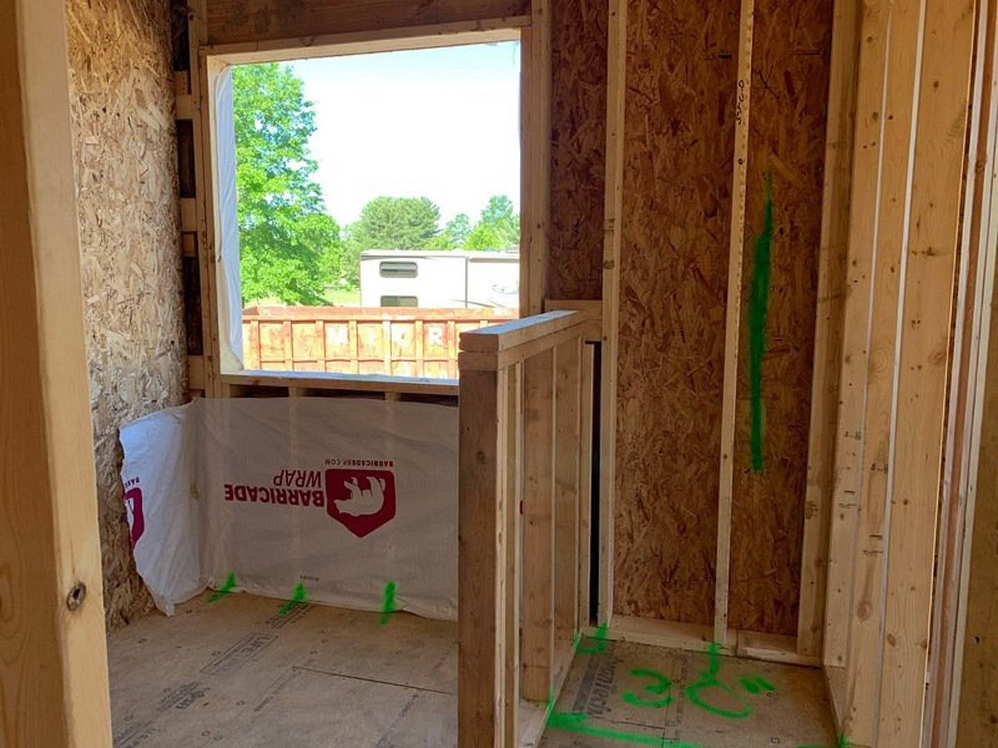 Unfinished room with exposed wood framing, white plastic wrap featuring red bear logo on wall, window overlooking trees and parked trailer, green paint markings on concrete floor