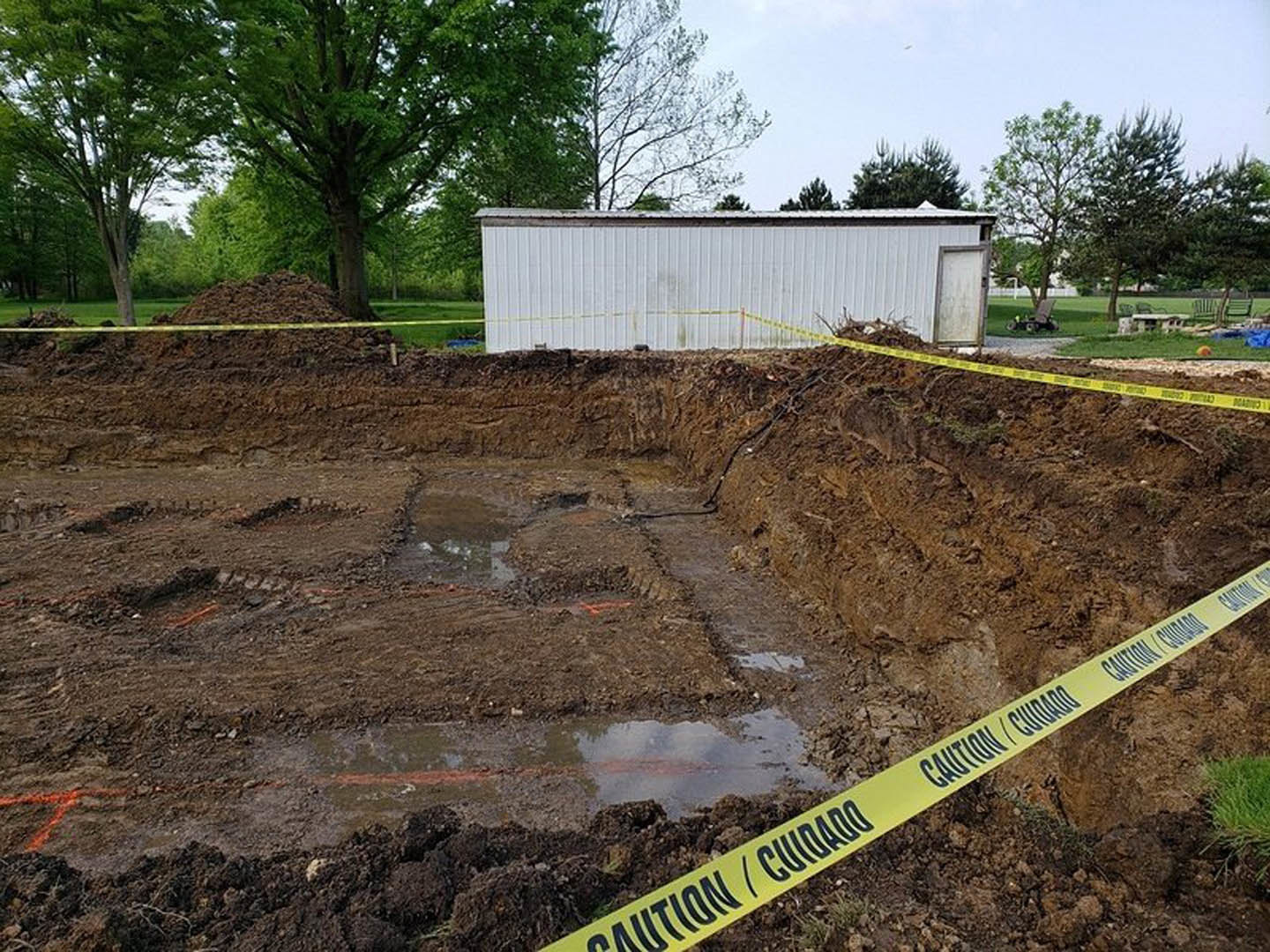 Yellow caution tape stretched across dirt lot beside white metal building, with pile of soil near tree and grassy area in foreground.