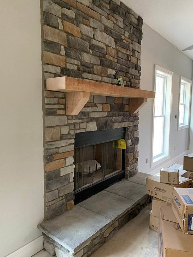 Stone fireplace with wood mantel, stone hearth step, window letting in natural light, wooden beam with paper, shelf, and hardwood flooring in a cozy room.