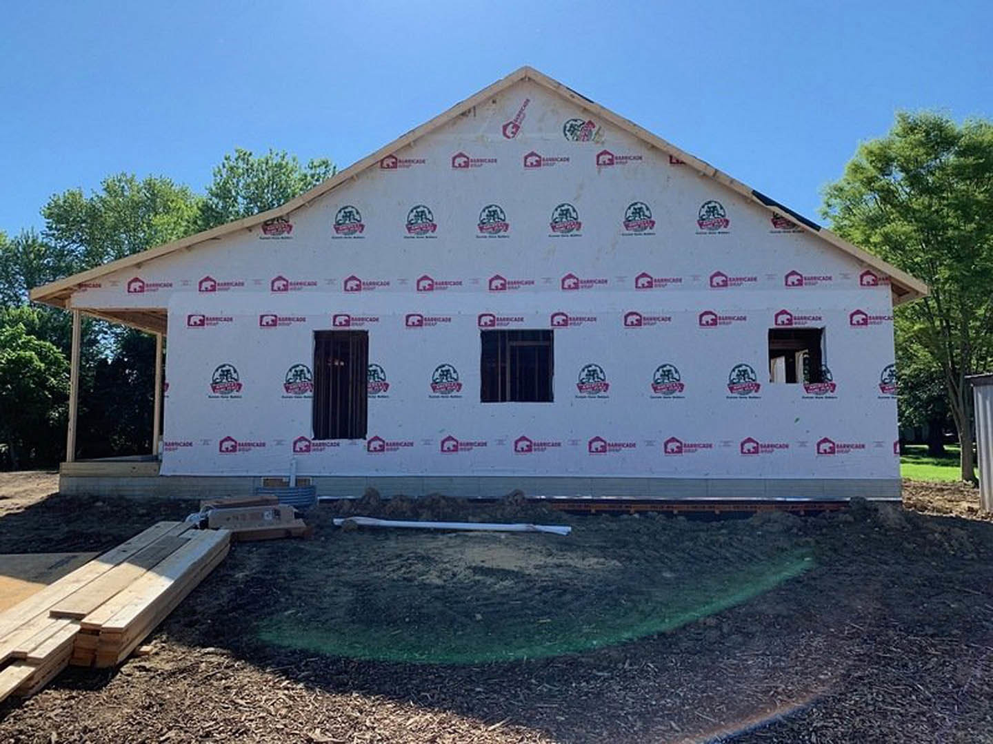 Wood-framed deck attached to a partially finished house with white exterior walls, exposed window, and stacked lumber on the ground