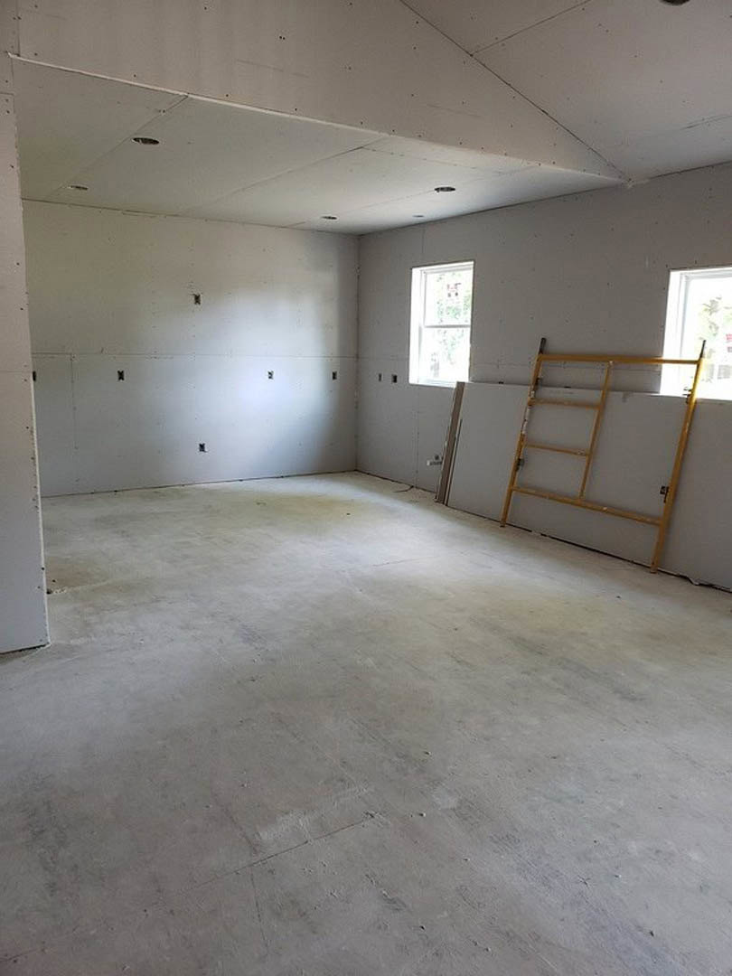 Unfinished room with plaster walls, white ceiling featuring recessed holes, ladder leaning against wall, window with white frame, and blurry chair in foreground