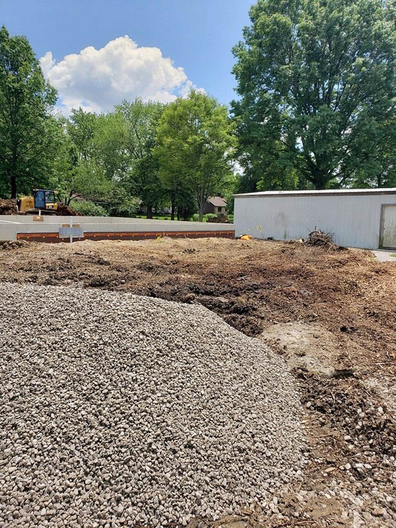 Gravel pile beside white exterior wall, dirt and rocks on ground, cloudy sky overhead, trees and plants in background