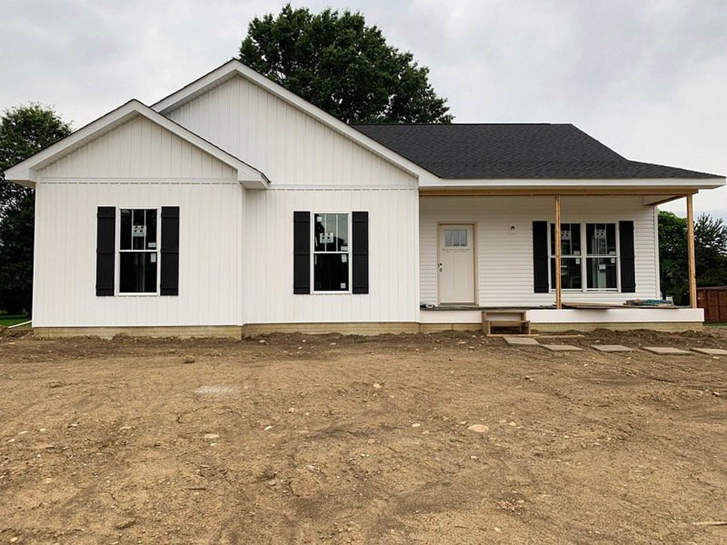 White siding house with black shutters, white front door with glass window, multiple windows with black trim, bare dirt landscaping, tree branches visible above roofline, clear sky