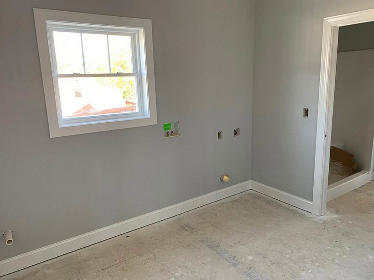 Bright room with white plaster walls, wood flooring, large window with white frame, white pipe running along wall, and white door; green sticker visible on wall near floor and