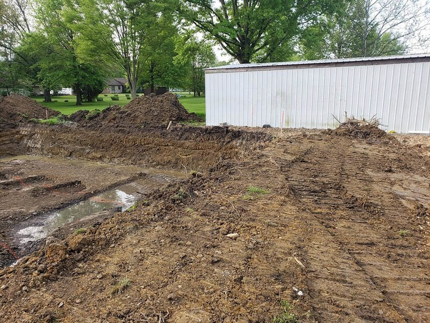 White metal shed surrounded by trees on a dirt field with scattered mud, wood pile, and puddle.