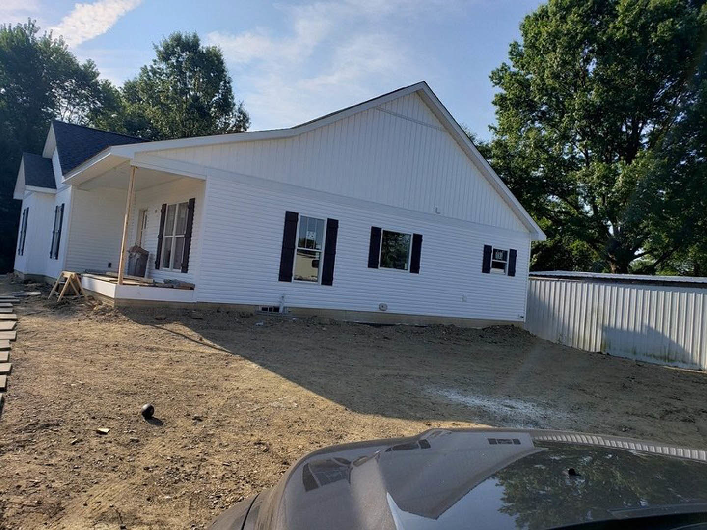 Two-story white house with black shutters under construction, surrounded by a wooden fence and mature trees, windows reflecting sky and greenery