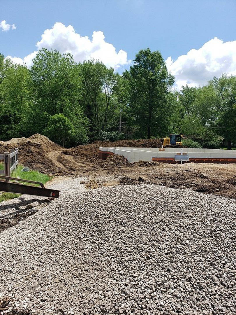 Gravel pile on bare soil at residential construction site, surrounded by trees under blue sky with scattered clouds