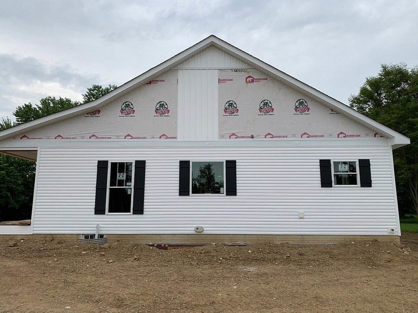 Partially built white house with black shutters, roof installed, windows visible, surrounded by dirt and construction materials, mature trees and historic Rockingham Meeting House
