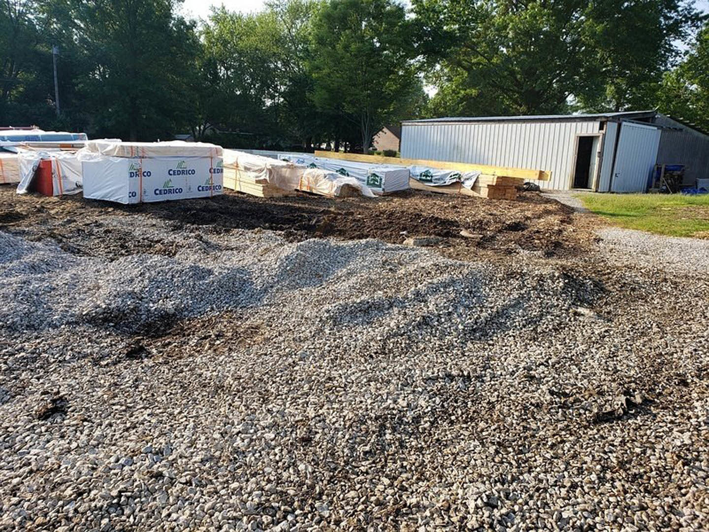 Gravel and wooden pallets stacked on bare ground beside a partially constructed home with open door, white utility box with blue writing, leafy tree, and close-up window visible