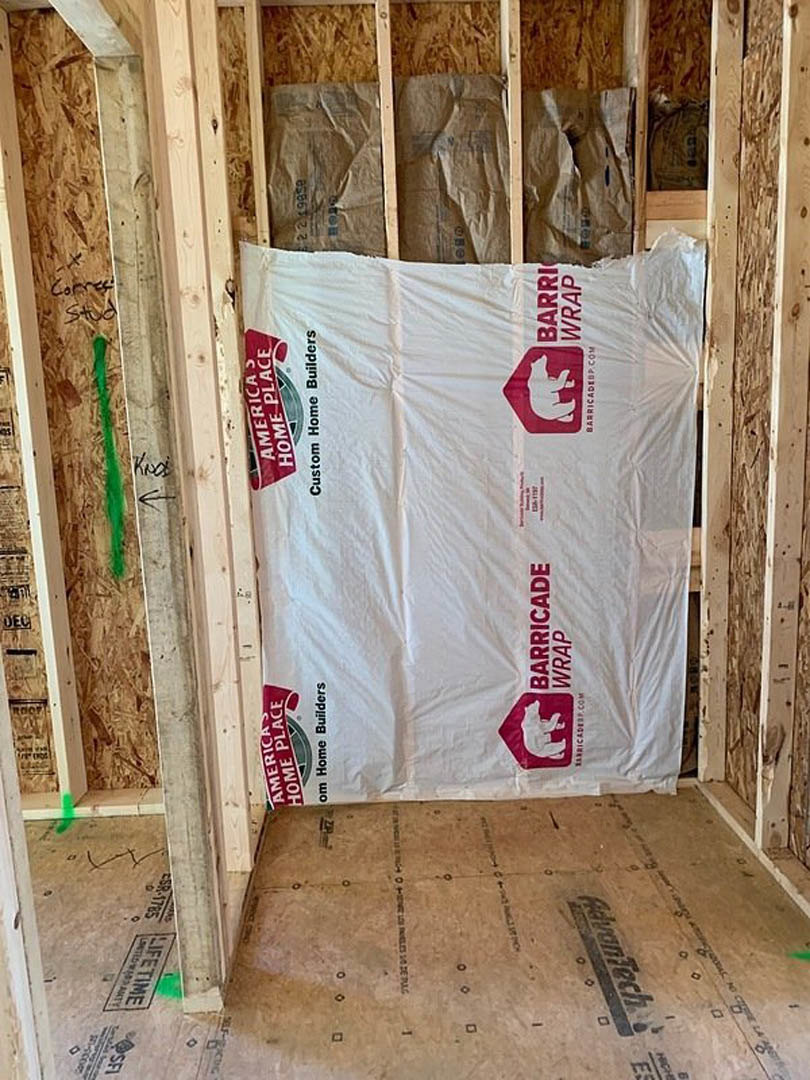 Wood-framed room under construction with exposed lumber, white plastic wrap featuring a red logo, and a red and white sign with a bear graphic.