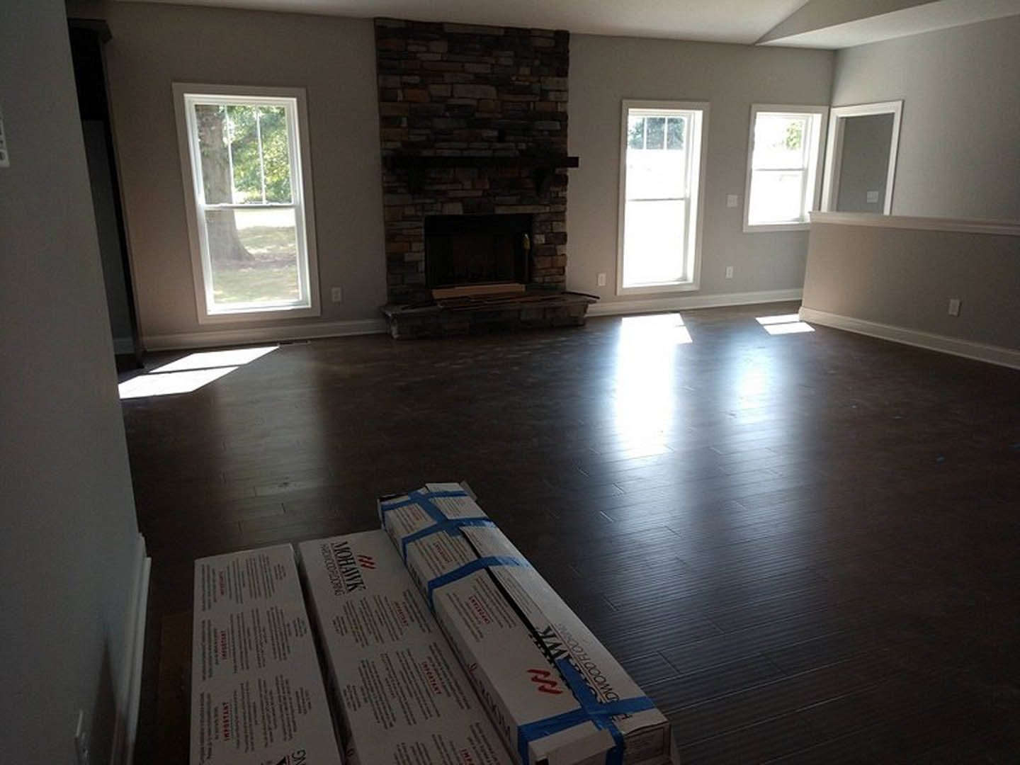 Living room with wood flooring, fireplace featuring a wood mantel, stack of moving boxes with blue tape, large window with white frame showing a tree outside