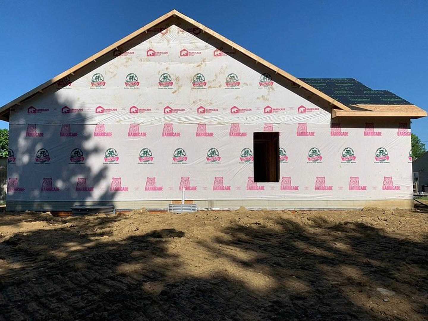 Partially built house with exposed framing, completed roof, and surrounding patch of bare dirt under clear sky