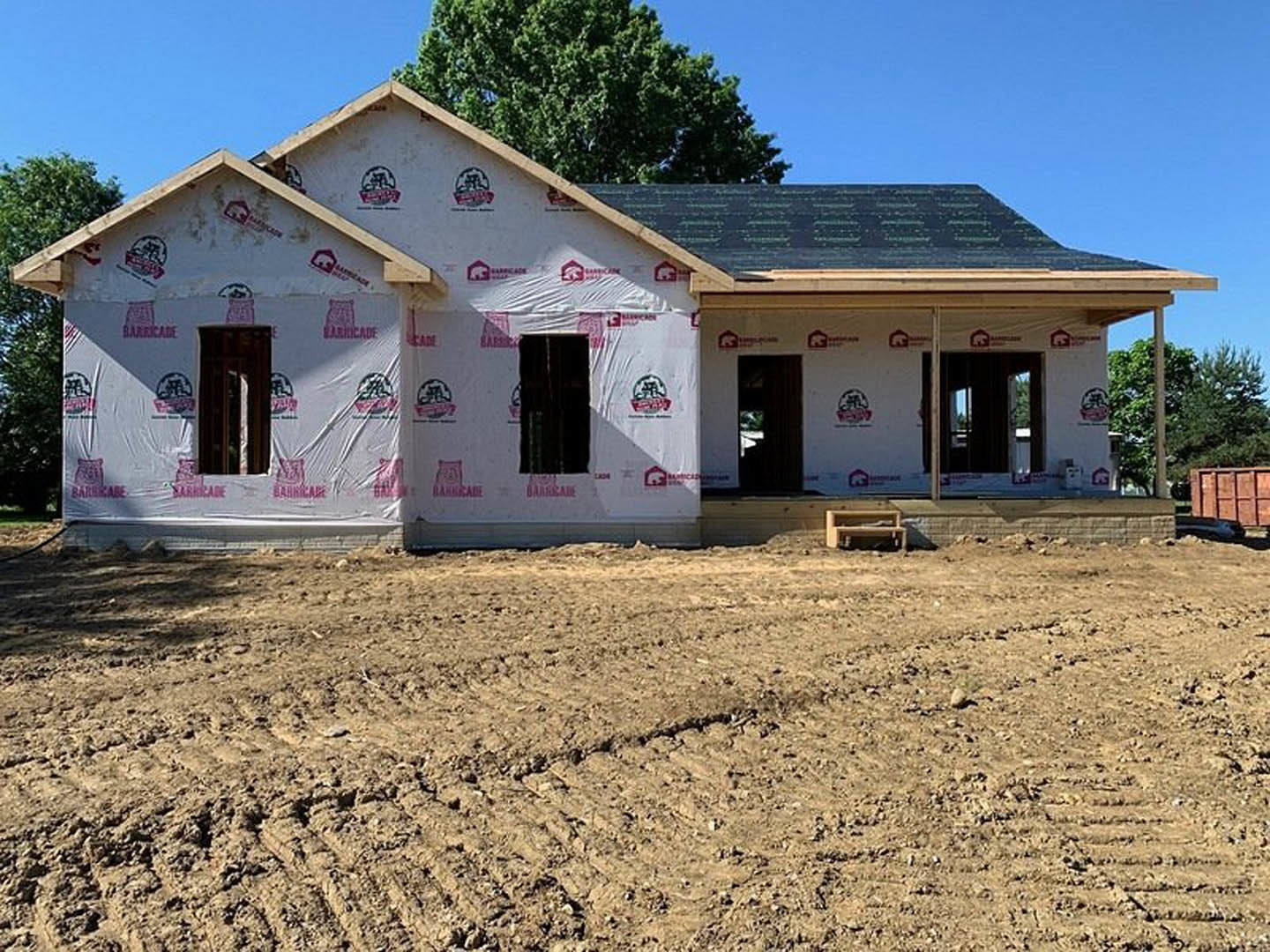Wood-framed house under construction on a dirt lot with a tree and open sky in the background