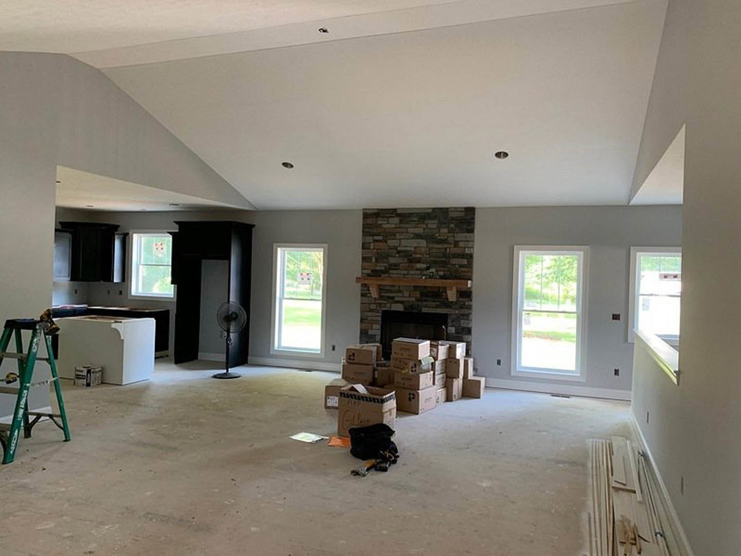 Living room with stacked moving boxes, stone fireplace, aluminum ladder with painter’s tape, ceiling fan, large windows showing outdoor view and a sign, light-colored plaster