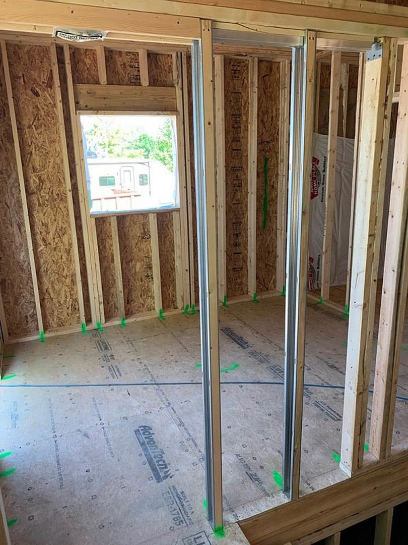 Room under construction with exposed wood framing, blue tape marking areas, window revealing white trailer outside, close-up of logo on grey surface