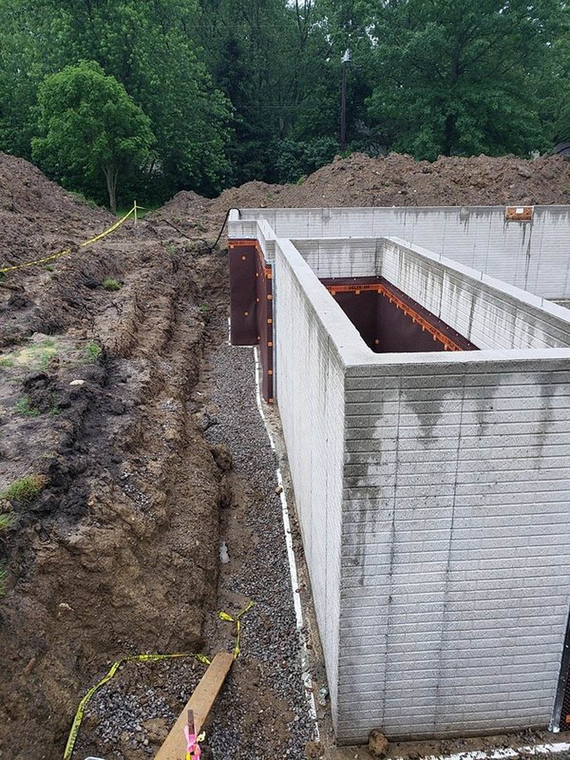 Concrete foundation wall with exposed metal beam, dirt piles, scattered rocks marked with yellow tape, and mature trees with green leaves surrounding the construction site