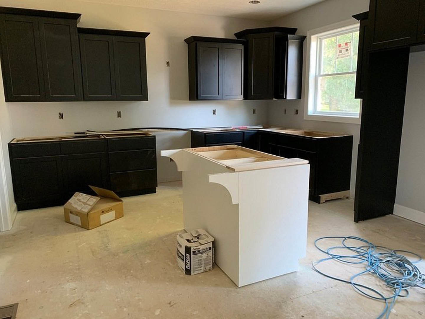 Kitchen with matte black cabinets, white quartz countertop, stainless steel sink, blue wire on light wood floor, white cabinet with cardboard box, window with posted sign