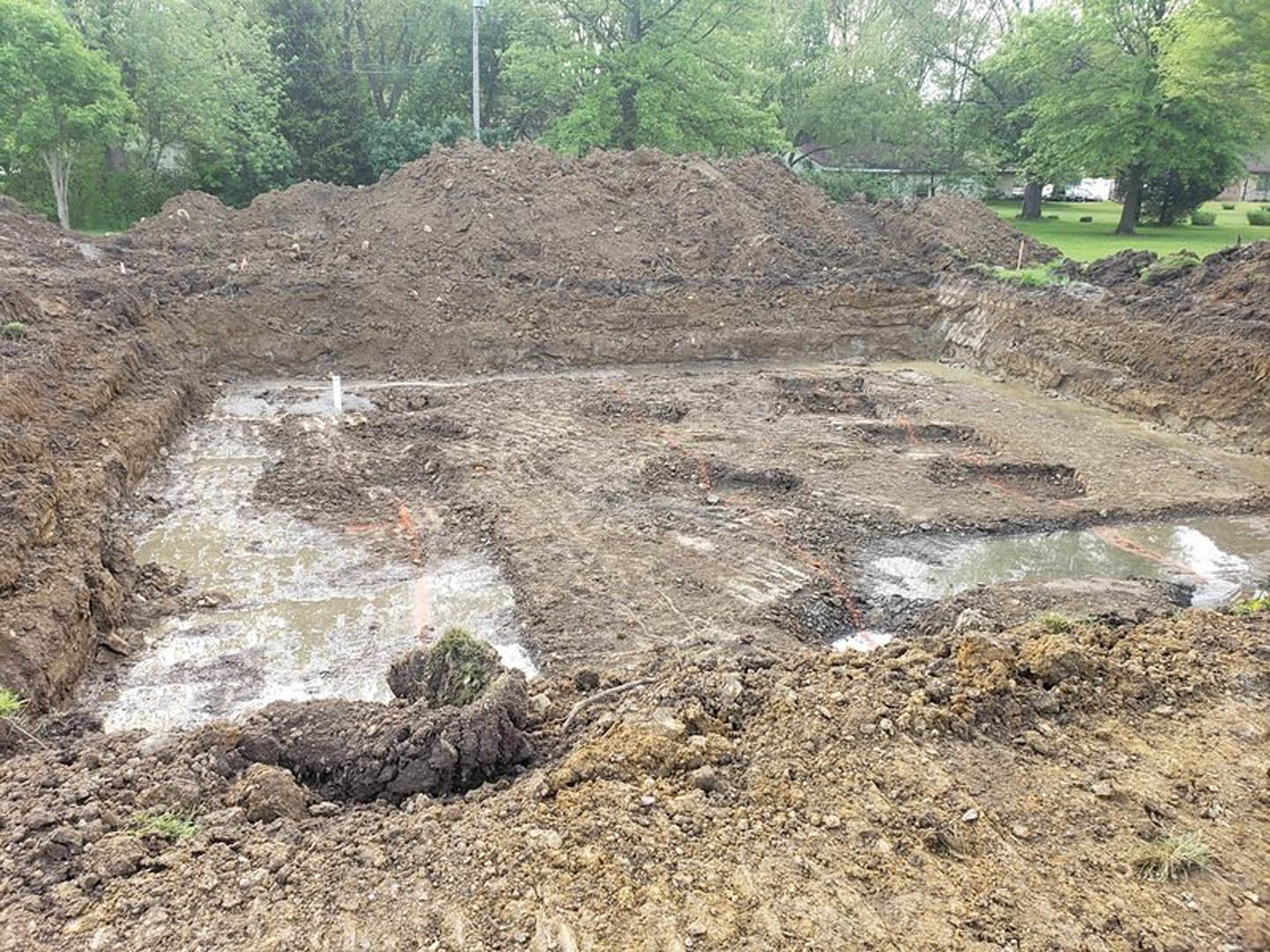 Expansive dirt excavation bordered by piles of soil and scattered trees, rough ground surface with exposed earth and rocks