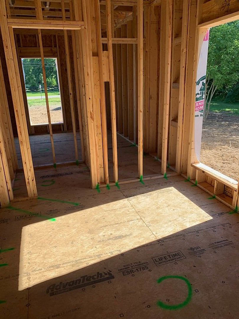 Room under construction with exposed wood framing, subfloor with green markings, window opening, and partial view of outdoor fence