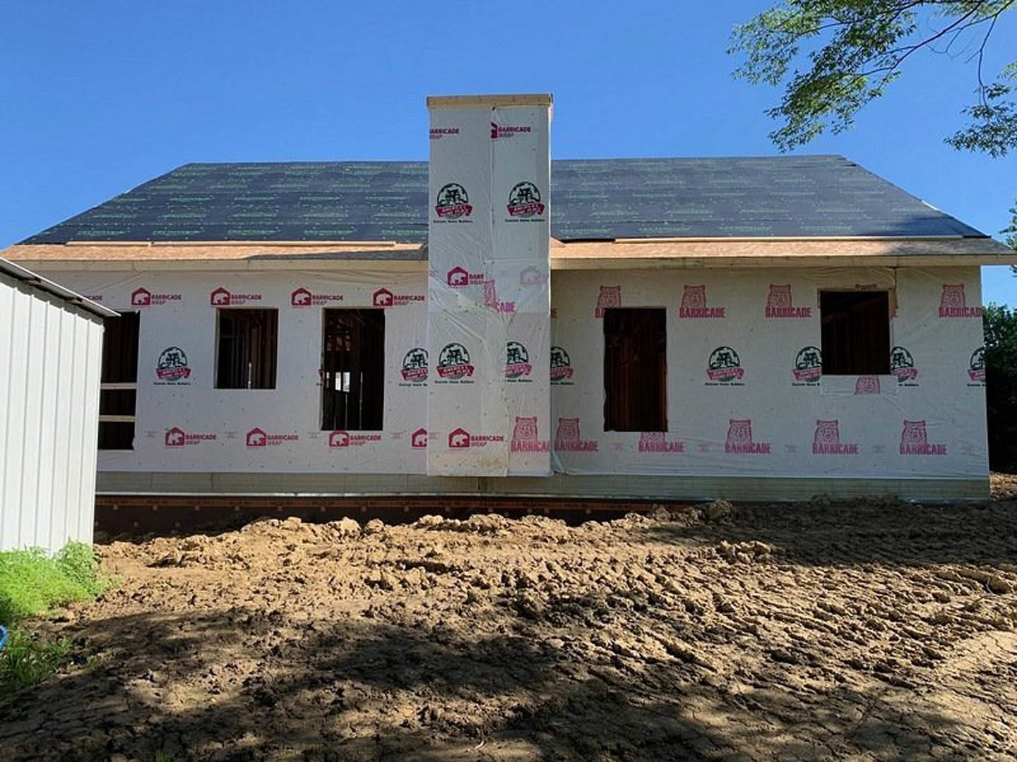 Framed house under construction with exposed insulation, brick chimney, white sign with red logo in front yard, pile of dirt on ground, unfinished siding, and windows