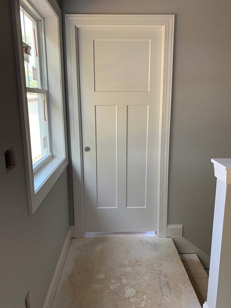 White paneled door with round silver knob in hallway, adjacent to white walls and baseboard molding, dirty beige floor, window with red tape visible in background.