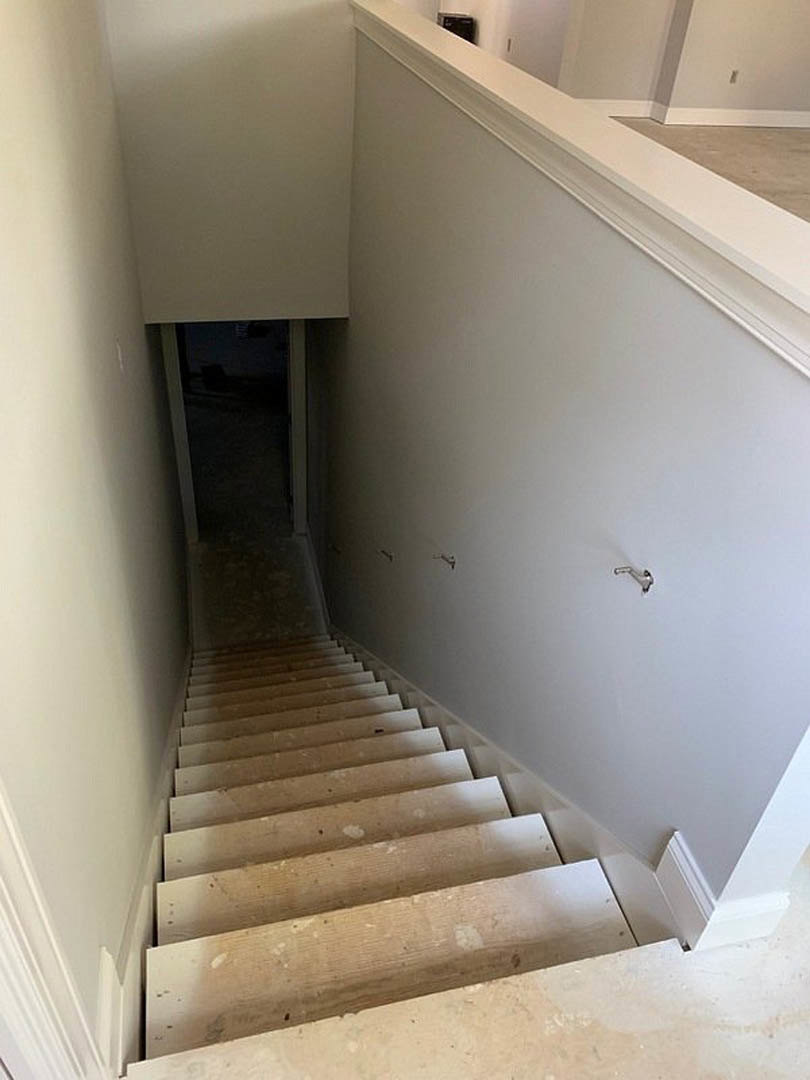 White staircase with wooden handrail ascending beside smooth plaster wall, red accent light, open doorway leading to dark hallway, composite flooring visible.
