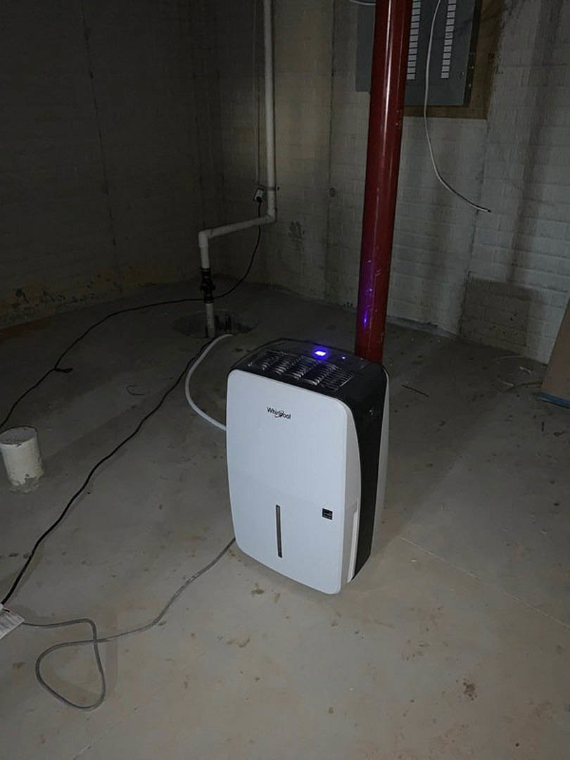 Wall-mounted white and black air conditioner with blue indicator light above a red pipe, set against a neutral interior wall and floor.
