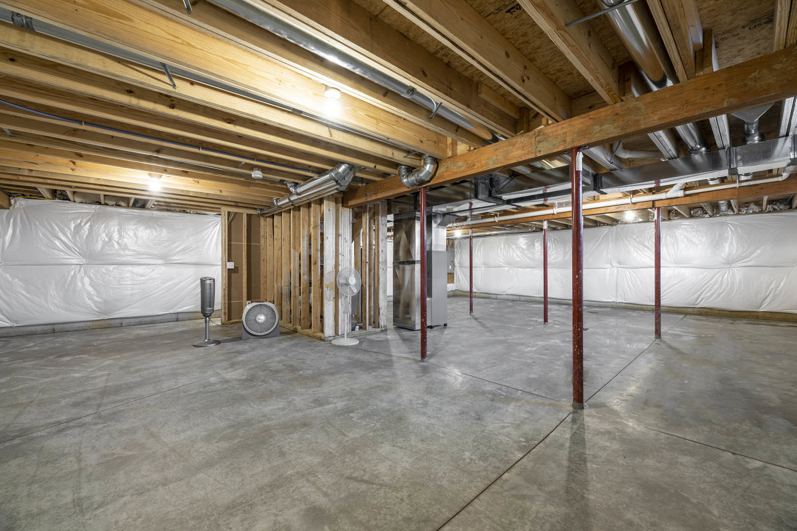 Exposed wood ceiling beams and metal pipes above a concrete floor, white plastic wall covering, steel support pole, white fan on wood surface