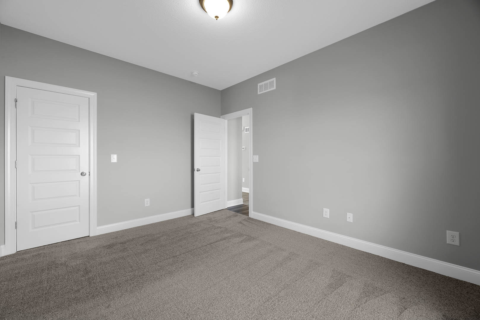 White paneled door with silver knob open to carpeted room, white walls with vent and electrical outlet, ceiling light fixture visible