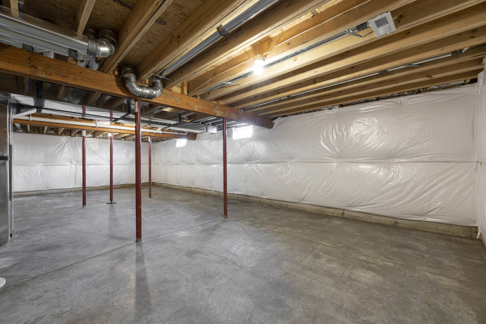 Basement with white plastic wall coverings, concrete floor, red support pole, exposed pipes and vents on ceiling, ceiling light fixture, wood beam with white vent