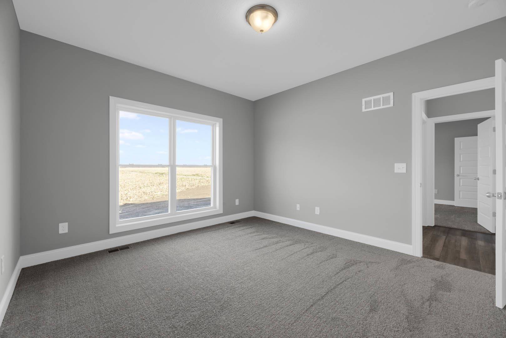 Carpeted room with white walls, large window overlooking grassy field, ceiling light fixture, white door with silver handles, and ceiling vent