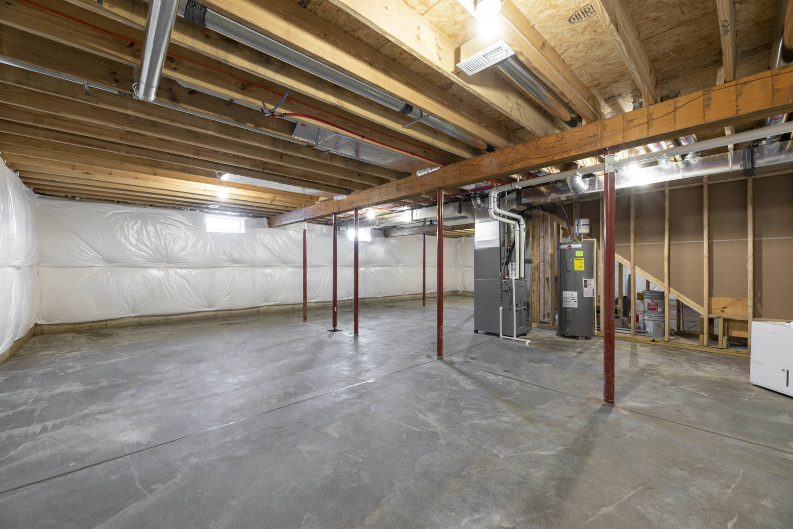 Basement room with exposed concrete floor, steel pipes, red support poles, grey container, white electrical box, and visible ceiling beams.
