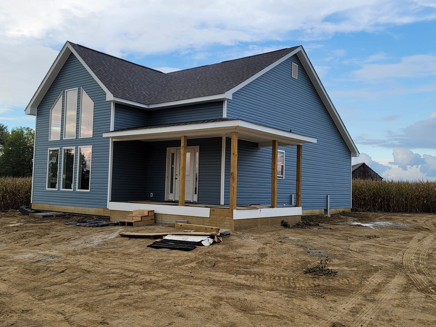 Partially built house with white siding, covered porch supported by wooden posts, white door with window, dirt yard scattered with lumber, tall grass field in background, cloudy