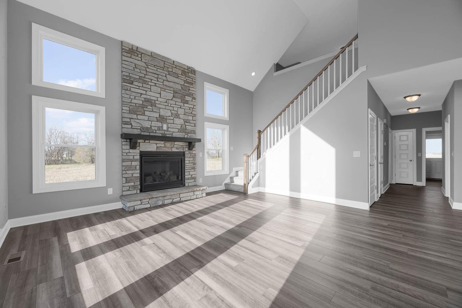Living room with wood flooring, glass-door fireplace with wood mantle, wooden staircase railing, large window overlooking field and trees