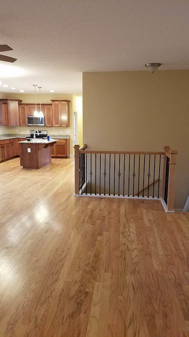 Open-concept kitchen and living room with wide plank wood floors, white cabinetry, marble countertop, black metal staircase railing, and ceiling fan