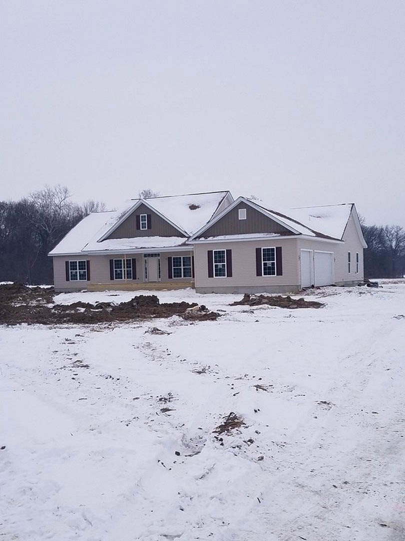 Two-story house with white trim and large windows, snow covering the roof and ground, bare trees nearby, overcast winter sky