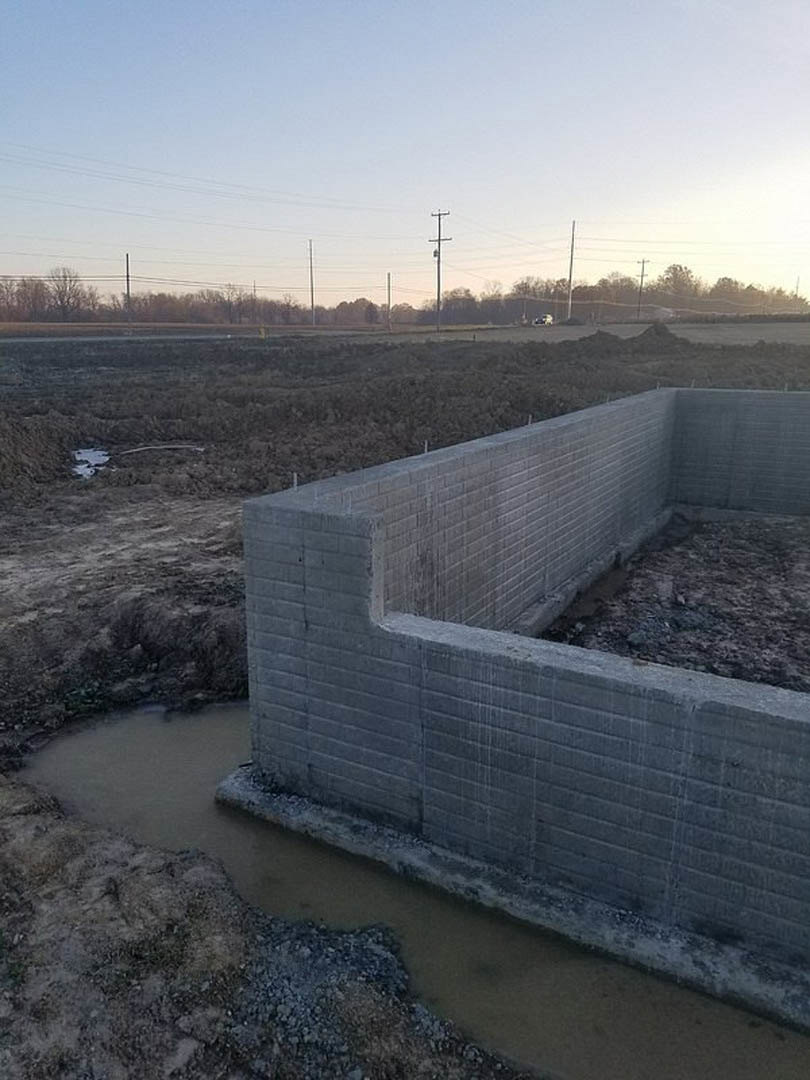 Concrete wall with water flowing through an opening, surrounded by rocky ground and dense bushes in a large field, telephone pole and power lines visible against the sky.