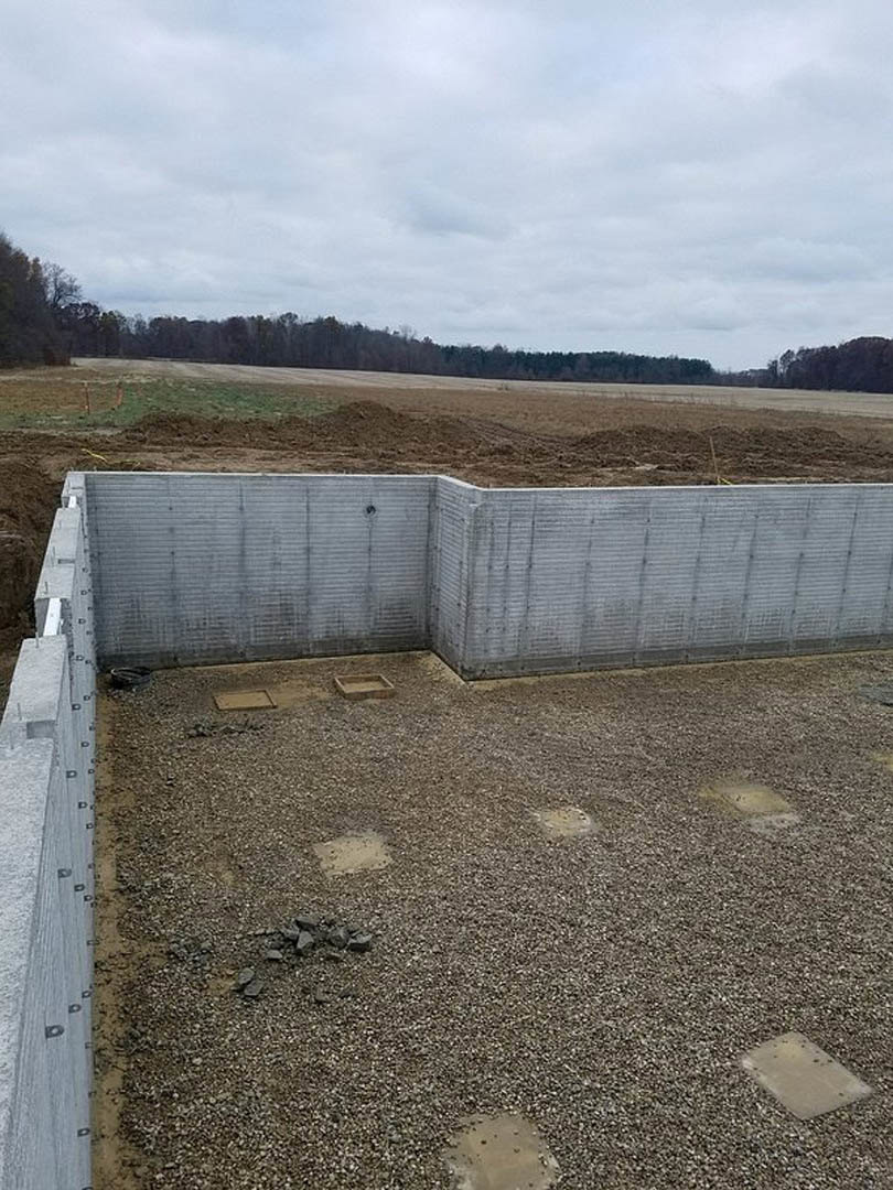 Concrete wall with a corner, gravel and rocks scattered on the ground, square object near a pile of rocks, cloudy sky above a forested area