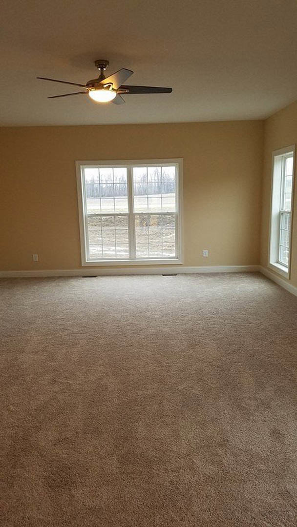 Beige carpeted room with white-framed windows, ceiling fan with light, plaster walls, and view of grassy field