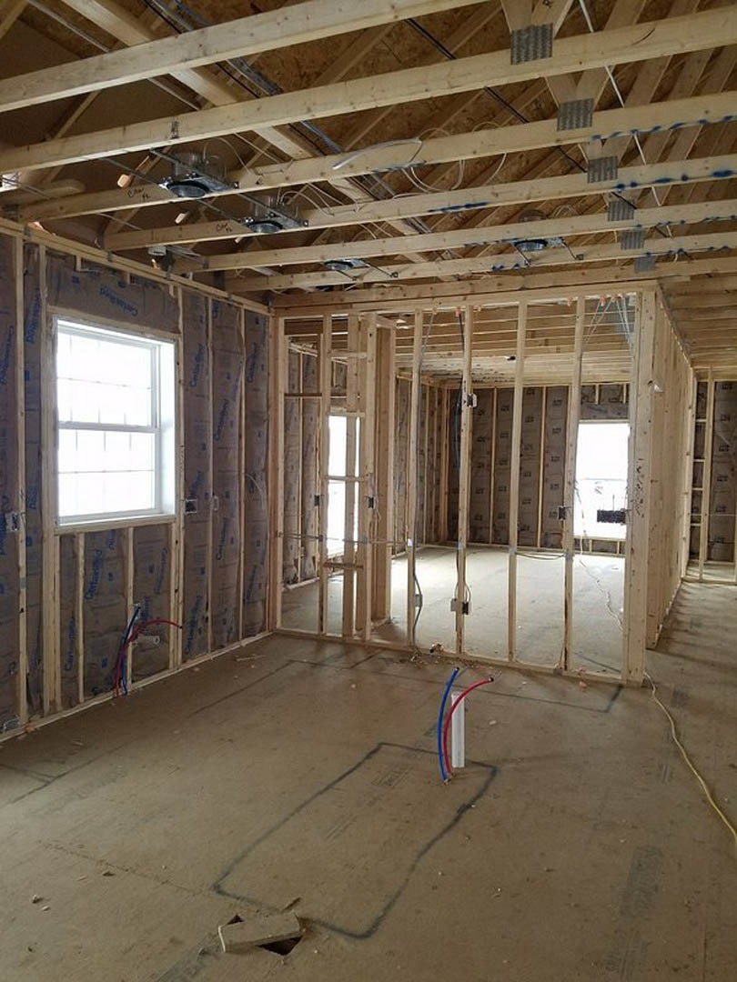 Wooden ceiling with exposed beams, white-framed window, unfinished floor with rectangular hole, red and blue tubing, construction materials visible.