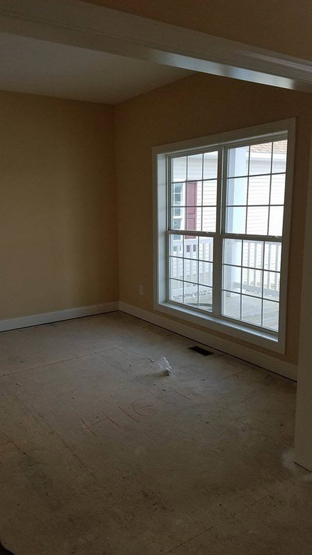 Sunlit room featuring a large window with white railing, smooth plaster walls, and concrete flooring with visible writing; deck area visible through the window.