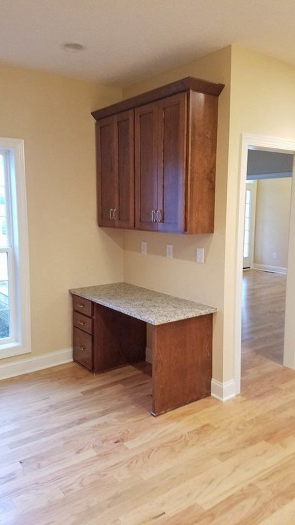 Marble-topped desk with drawers set on hardwood floor beside white-framed window, surrounded by light wood cabinetry and neutral walls