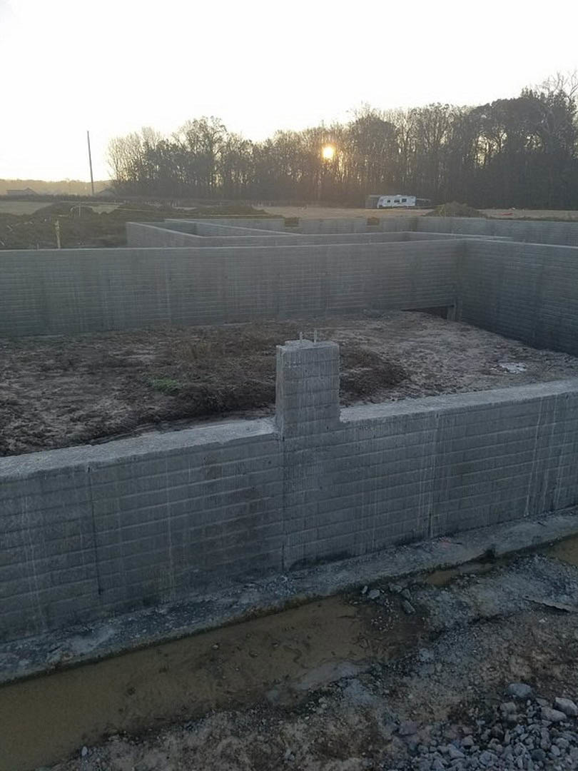 Concrete foundation with exposed hole in the ground, surrounded by dirt and water, sunlight filtering through nearby trees, grey concrete walls featuring square patterns and