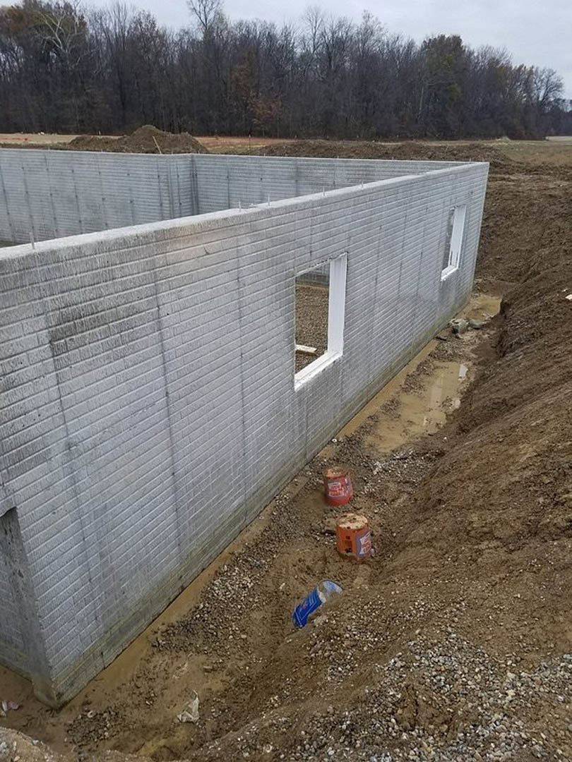 Partially built house with exposed foundation, dirt excavation in foreground, unfinished exterior walls, surrounding soil and scattered trees under open sky